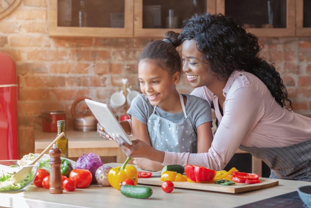 African American mother and daughter look at tablet and prepare to make dinner with vegetables on a cutting board in front of them.
