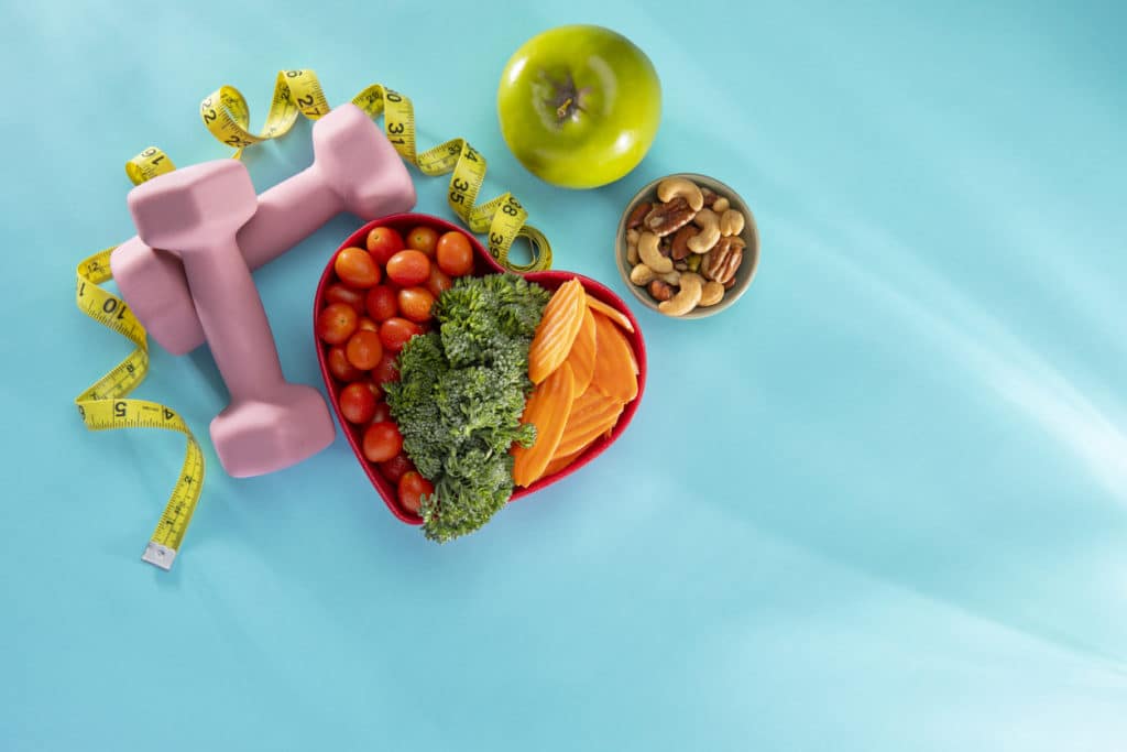 Vegetables in a heart bowl with dumbells and an apple on a blue background