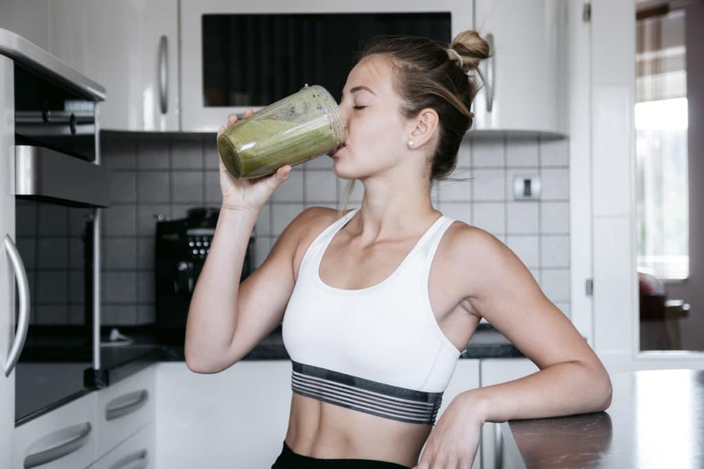 Young fit woman wearing fitness clothes drinks a green smoothie in her kitchen.