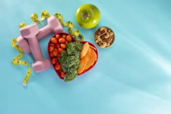Vegetables in a heart bowl with dumbells and an apple on a blue background