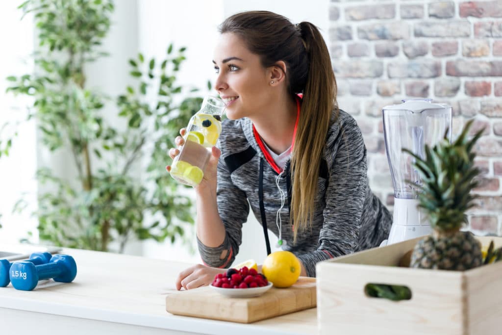 Mulher jovem desportiva olhando de soslaio enquanto bebe suco de limão na cozinha em casa.