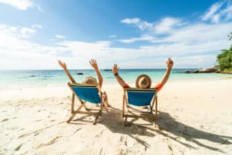 Two happy people sit in chairs on the beach, prioritizing taking time off.