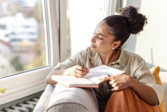 A woman gazes out the window as she writes in her gratitude journal to improve physical and mental health.